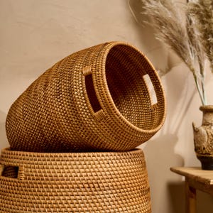 Op de afbeelding: Twee ronde, gevlochten rotan manden verticaal op elkaar gestapeld. De bovenste mand staat in een hoek, waardoor de holle binnenkant en de geïntegreerde handgrepen aan de zijkant zichtbaar zijn. Beide manden hebben een lichtbruin vlechtwerk.