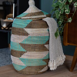 Puede incluir: Una cesta de almacenamiento grande y tejida con tapa. La cesta est&aacute; hecha de materiales naturales y tiene un patr&oacute;n geom&eacute;trico en tonos marrones, blancos y turquesa. Una manta de lana gris est&aacute; drapeada sobre el lado de la cesta.