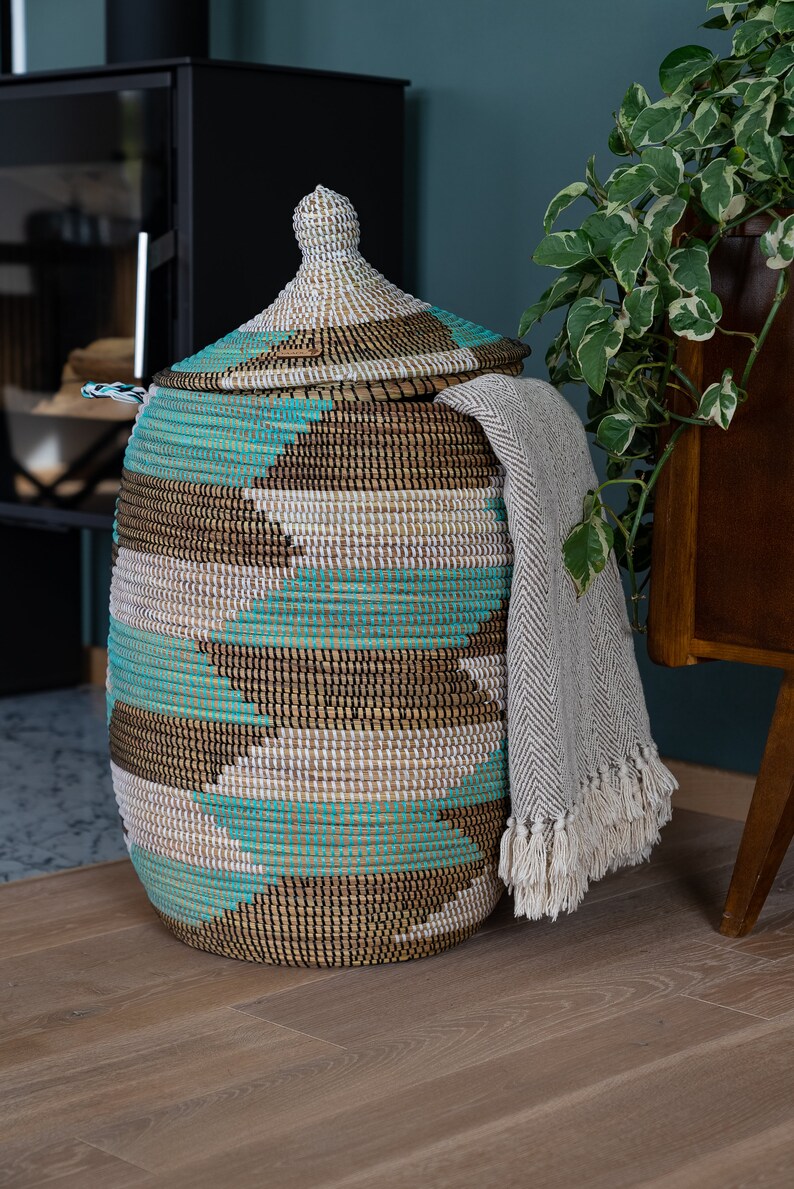 Puede incluir: Una cesta de almacenamiento grande y tejida con tapa. La cesta est&aacute; hecha de materiales naturales y tiene un patr&oacute;n geom&eacute;trico en tonos marrones, blancos y turquesa. Una manta de lana gris est&aacute; drapeada sobre el lado de la cesta.