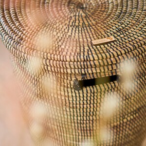 May include: A woven storage basket with a black and natural straw pattern. The basket has a lid and a small opening on the side. The basket has a wooden tag with the word "YAADU" on it.