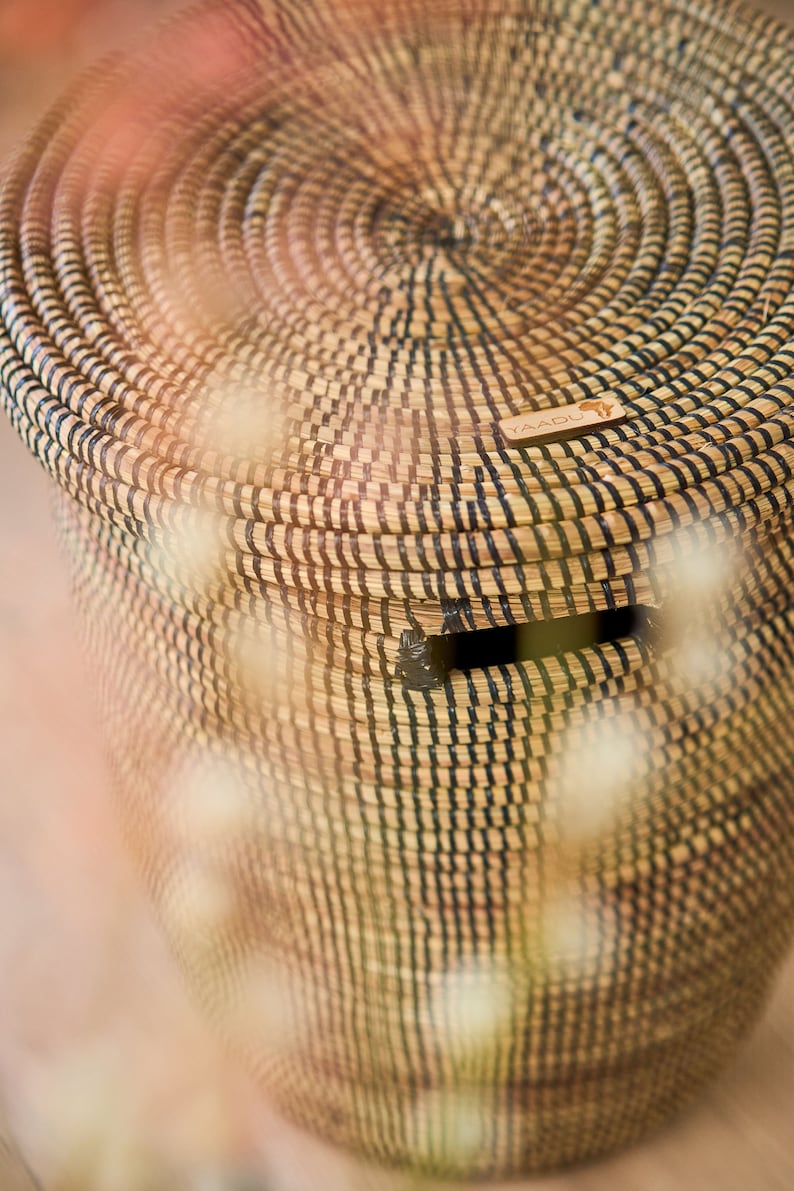 May include: A woven storage basket with a black and natural straw pattern. The basket has a lid and a small opening on the side. The basket has a wooden tag with the word "YAADU" on it.