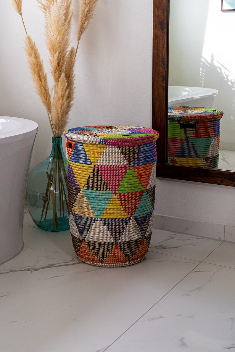 Puede incluir: Una cesta de almacenamiento redonda tejida con tapa. La cesta presenta un patr&oacute;n geom&eacute;trico de tri&aacute;ngulos coloridos en tonos de amarillo, rosa, azul y negro. La cesta est&aacute; en un ba&ntilde;o.