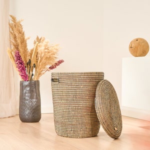 May include: A woven storage basket with a lid, made of natural materials, with a dark brown and beige pattern. The basket is open and the lid is sitting next to it on a light wood floor.
