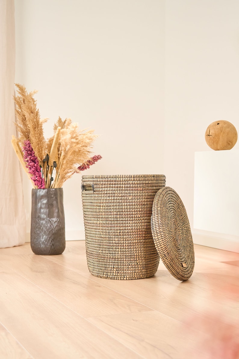 May include: A woven storage basket with a lid, made of natural materials, with a dark brown and beige pattern. The basket is open and the lid is sitting next to it on a light wood floor.