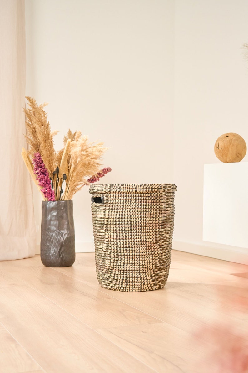 May include: A woven black and tan storage basket with a black handle. The basket is sitting on a light wood floor.
