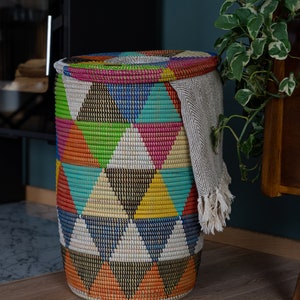 Puede incluir: Una cesta redonda tejida con tapa, con un colorido patr&oacute;n geom&eacute;trico de tri&aacute;ngulos en tonos de azul, verde, naranja y rosa. Una manta de color claro con flecos est&aacute; drapeada sobre el lateral. La cesta est&aacute; sobre un suelo de madera.