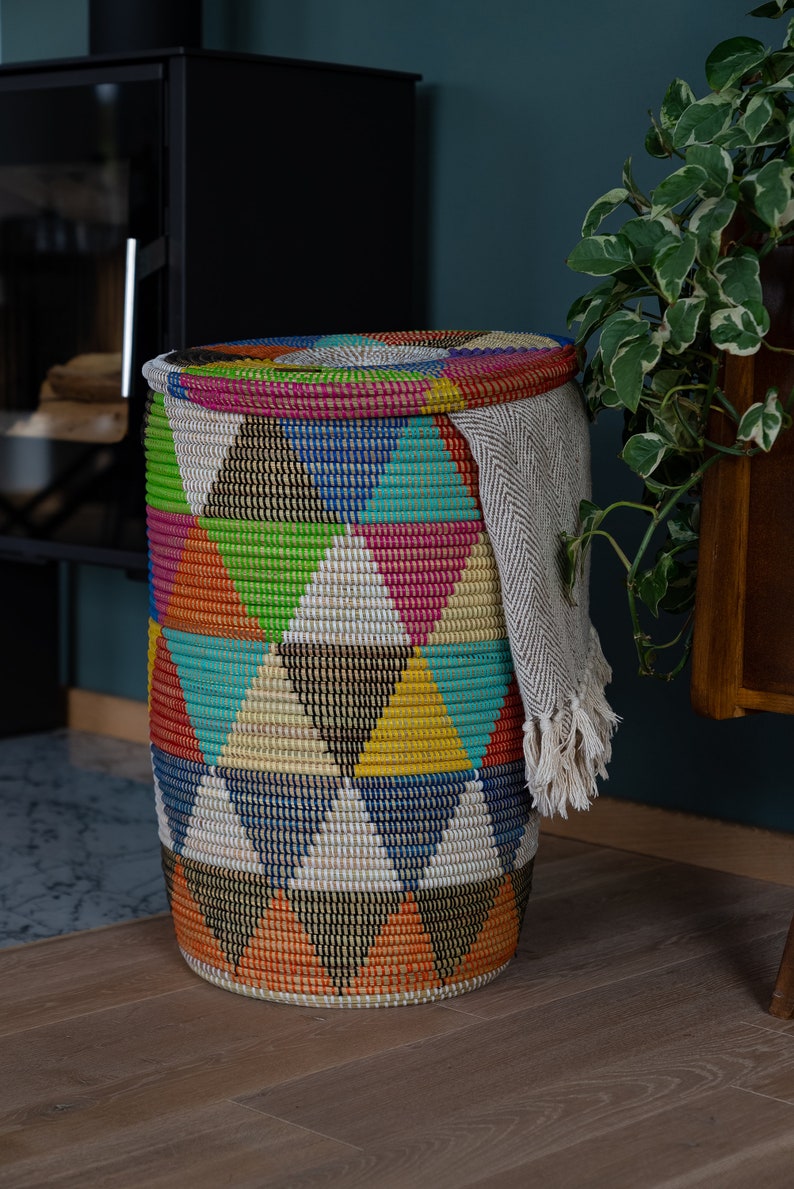 Puede incluir: Una cesta redonda tejida con tapa, con un colorido patr&oacute;n geom&eacute;trico de tri&aacute;ngulos en tonos de azul, verde, naranja y rosa. Una manta de color claro con flecos est&aacute; drapeada sobre el lateral. La cesta est&aacute; sobre un suelo de madera.