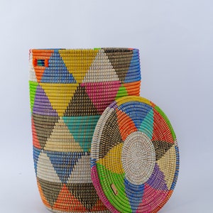 Puede incluir: Una cesta tejida cil&iacute;ndrica con tapa, con un vibrante patr&oacute;n geom&eacute;trico. El dise&ntilde;o de la cesta incorpora tri&aacute;ngulos y c&iacute;rculos conc&eacute;ntricos en un espectro de colores, incluyendo azul, amarillo, rosa y naranja. La tapa es redonda.