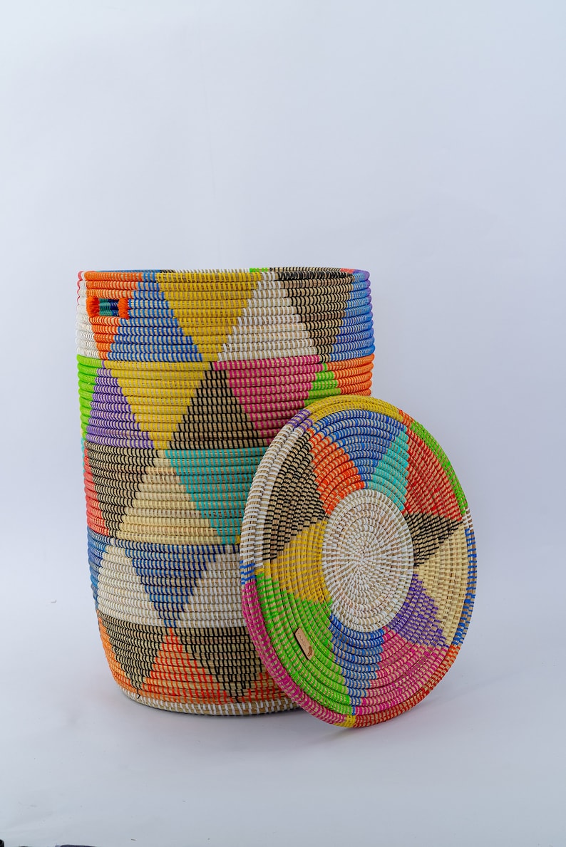 Puede incluir: Una cesta tejida cil&iacute;ndrica con tapa, con un vibrante patr&oacute;n geom&eacute;trico. El dise&ntilde;o de la cesta incorpora tri&aacute;ngulos y c&iacute;rculos conc&eacute;ntricos en un espectro de colores, incluyendo azul, amarillo, rosa y naranja. La tapa es redonda.