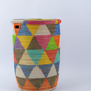 Puede incluir: Una cesta alta y cil&iacute;ndrica tejida con tapa. La cesta presenta un vibrante patr&oacute;n geom&eacute;trico de tri&aacute;ngulos en varios colores, incluyendo rosa, amarillo, azul y naranja. La tapa tiene una combinaci&oacute;n de colores a juego. La cesta probablemente se utiliza para el almacenamiento.