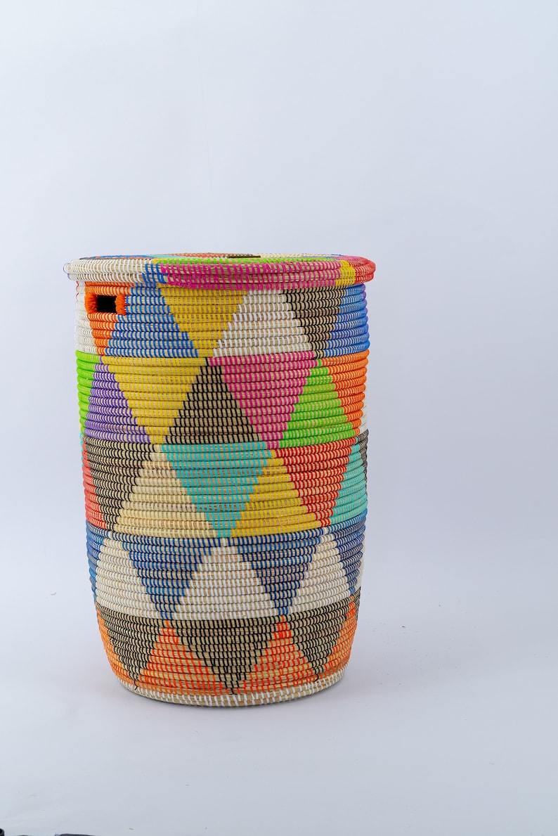 Puede incluir: Una cesta alta y cil&iacute;ndrica tejida con tapa. La cesta presenta un vibrante patr&oacute;n geom&eacute;trico de tri&aacute;ngulos en varios colores, incluyendo rosa, amarillo, azul y naranja. La tapa tiene una combinaci&oacute;n de colores a juego. La cesta probablemente se utiliza para el almacenamiento.