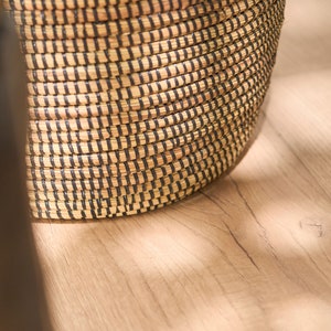 May include: A woven basket with black and natural brown stripes. The basket is made of natural materials and has a round shape.