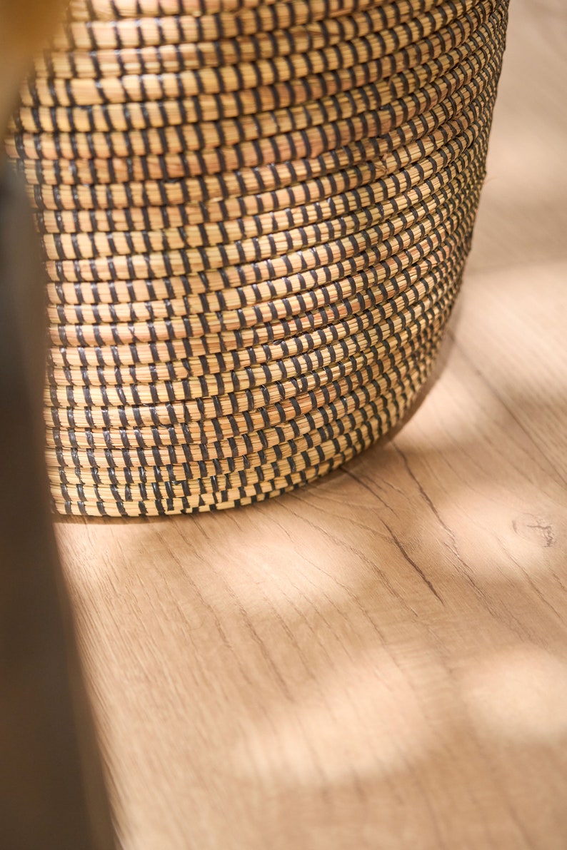 May include: A woven basket with black and natural brown stripes. The basket is made of natural materials and has a round shape.