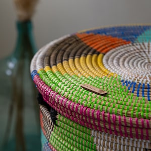 Puede incluir: Una cesta redonda y tejida con tapa, con una vibrante gama de colores que incluyen verde, rosa, amarillo, naranja y azul. La cesta tiene un aspecto natural y texturizado con una peque&ntilde;a etiqueta de madera. Un jarr&oacute;n de cristal turquesa est&aacute; borroso en el fondo.