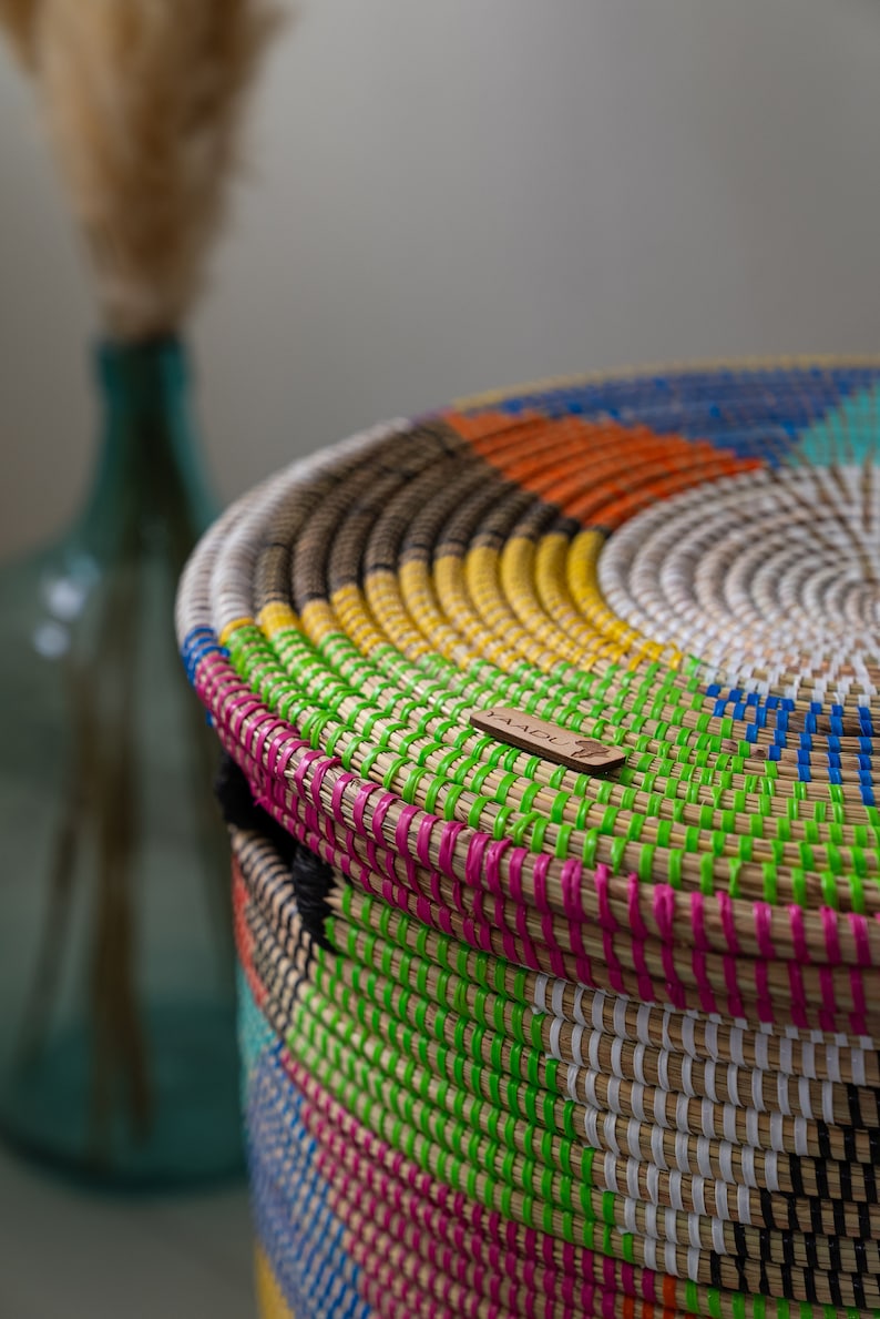 Puede incluir: Una cesta redonda y tejida con tapa, con una vibrante gama de colores que incluyen verde, rosa, amarillo, naranja y azul. La cesta tiene un aspecto natural y texturizado con una peque&ntilde;a etiqueta de madera. Un jarr&oacute;n de cristal turquesa est&aacute; borroso en el fondo.
