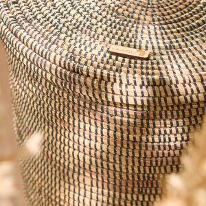 May include: A large, woven basket with black and natural colored stripes. The basket is made of natural materials and has a round shape. The basket has a wooden tag with the word "YAADU" on it.
