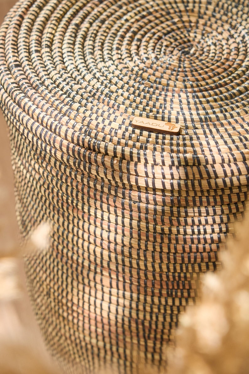 May include: A large, woven basket with black and natural colored stripes. The basket is made of natural materials and has a round shape. The basket has a wooden tag with the word "YAADU" on it.