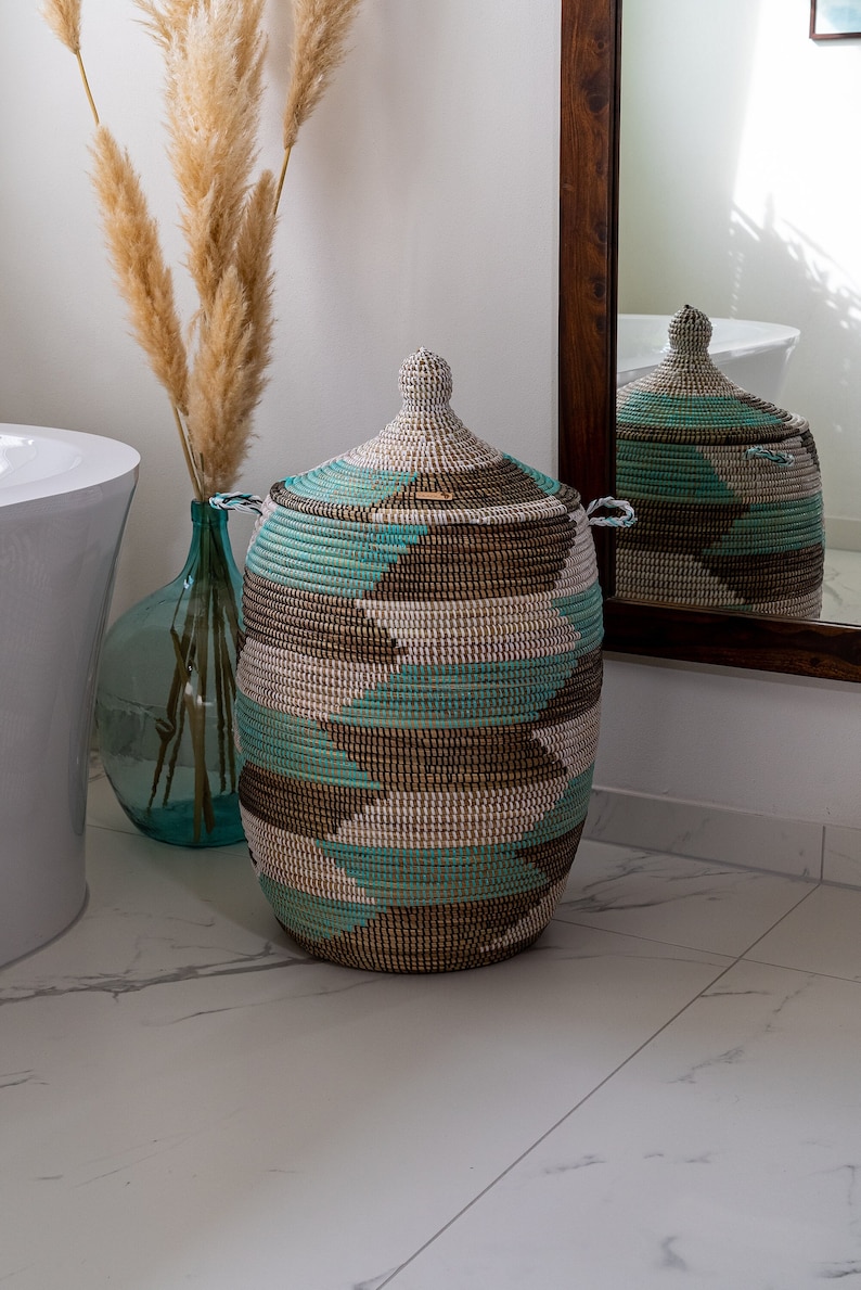 Puede incluir: Una cesta de almacenamiento grande y tejida con tapa. La cesta est&aacute; hecha de materiales naturales y tiene un patr&oacute;n geom&eacute;trico en tonos marrones, blancos y azul verdoso.