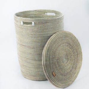 K&ouml;nnte beinhalten: Ein hoher, zylindrischer W&auml;schekorb mit passendem Deckel. Der Korb ist aus Naturfasern in Beige-, Wei&szlig;- und Gr&uuml;nt&ouml;nen gewebt. Der runde Deckel ist teilweise ge&ouml;ffnet. Der Korb hat zwei Griffe und ein kleines Lederetikett.