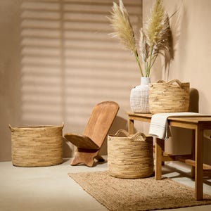 Op de afbeelding: Twee gevlochten strooien manden met handvatten, een houten stoel, een witte, getextureerde vaas met hoog gedroogd gras en een houten bank met een witte doek. De manden zijn rond en ovaal, met een natuurlijke beige kleur en een gewikkelde structuur.