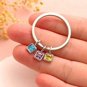 May include: A silver keychain with three square charms in blue, purple, and yellow. Each charm has a gemstone set in a silver frame. The keychain is held in a hand, with a soft pink background.