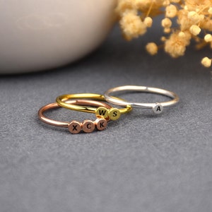 Op de afbeelding: Drie delicate ringen met initial charms. De ringen zijn gestapeld op een grijs oppervlak. De bovenste ring is zilver met de letter "A" charm. De middelste ring is goud met de letters "W" en "S" charms. De onderste ring is rosé goud met de letters "X", "C", "K" charms.