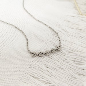 May include: A silver chain necklace with a pendant that spells out the word "BRAVE" in individual silver circles.