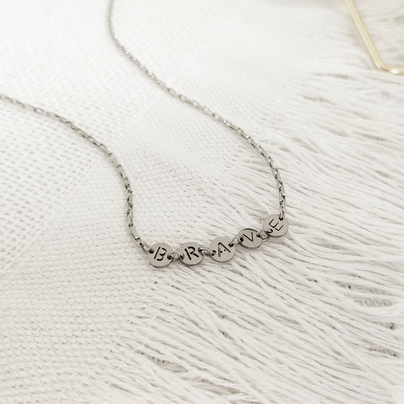 May include: A silver chain necklace with a pendant that spells out the word "BRAVE" in individual silver circles.