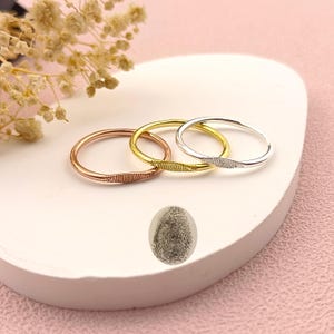 Può includere: Tre sottili anelli in oro rosa, oro e argento, ciascuno con un motivo a impronta digitale. Gli anelli sono esposti su una superficie bianca con uno sfondo rosa. Viene mostrata anche un'immagine dell'impronta digitale.
