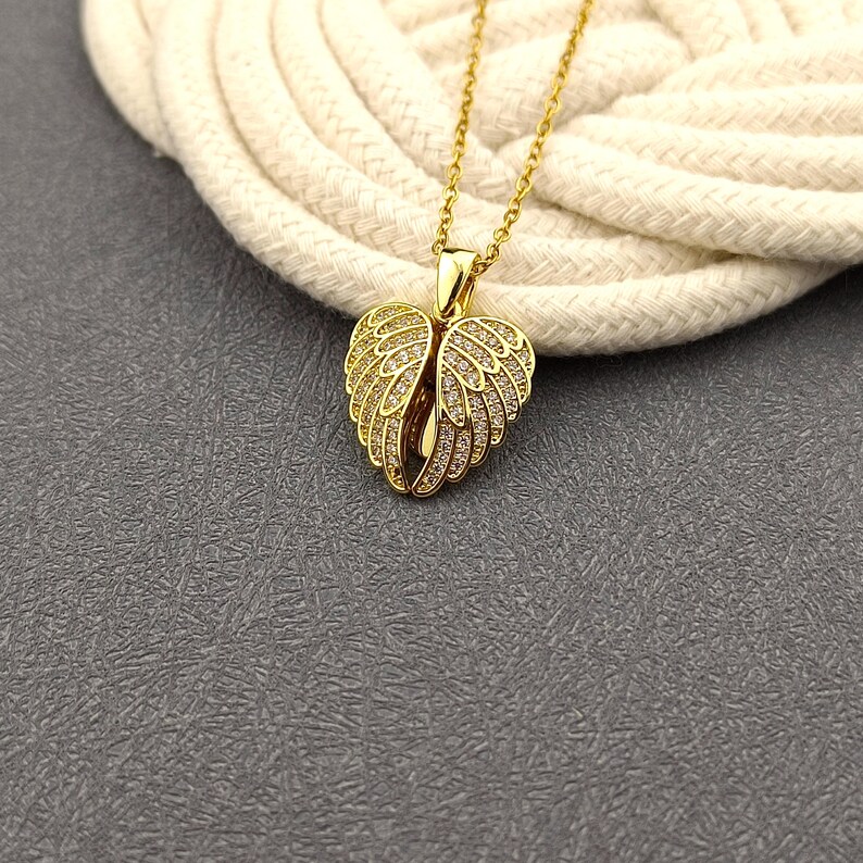May include: Gold necklace with a heart-shaped pendant featuring angel wings adorned with sparkling clear stones.