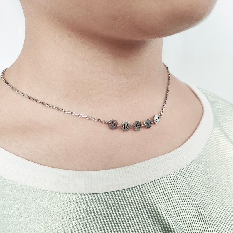 May include: A silver chain necklace with a pendant that spells out the word "BRAVE" in individual silver circles.