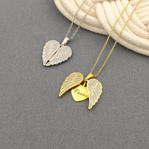 Op de afbeelding: Twee gouden kettingen met engelenvleugel hangers. Een hanger is een hart met het woord "Angel" erop gegraveerd. De andere hanger is een paar engelenvleugels.