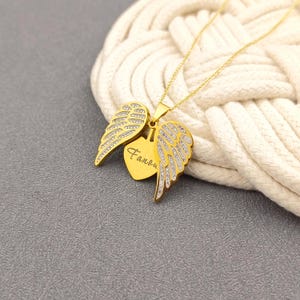 K&ouml;nnte beinhalten: Goldkette mit einem Herzanh&auml;nger mit dem Wort "Fanou" und zwei Engelsfl&uuml;geln mit funkelnden Akzenten.