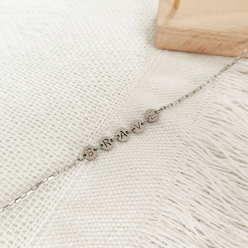 May include: A silver chain necklace with round silver charms spelling out the word "BRAVE".