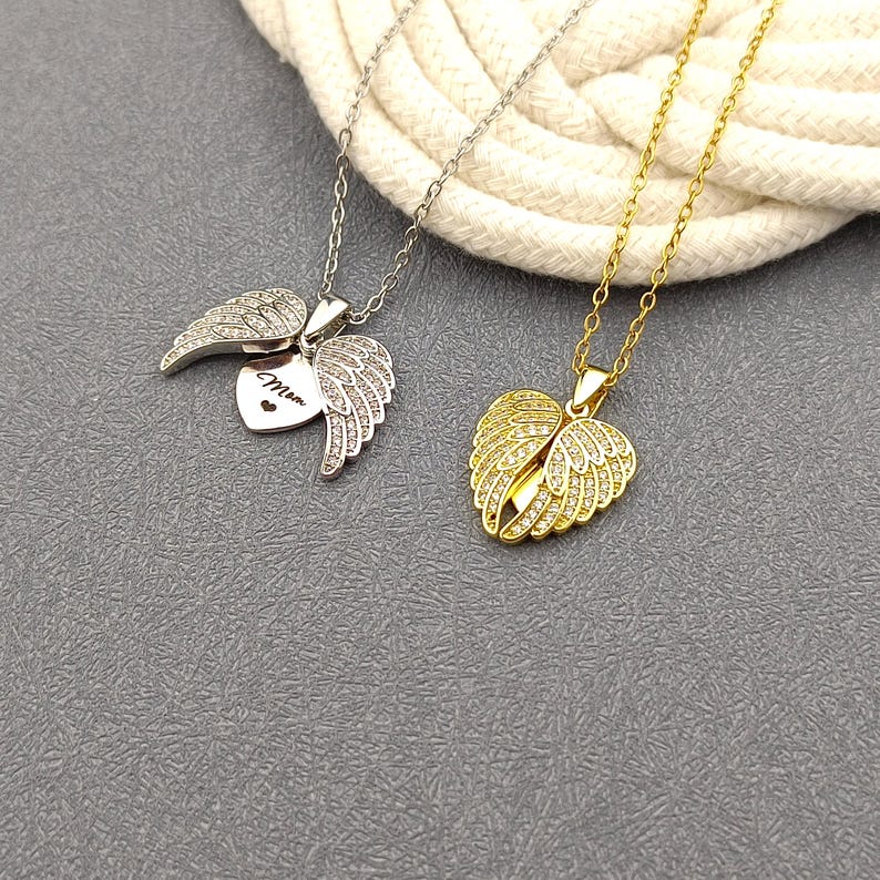 May include: Two silver and gold necklaces with angel wing pendants. The silver pendant has a heart with the word "Mom" inscribed on it. Both pendants are adorned with clear cubic zirconia stones.