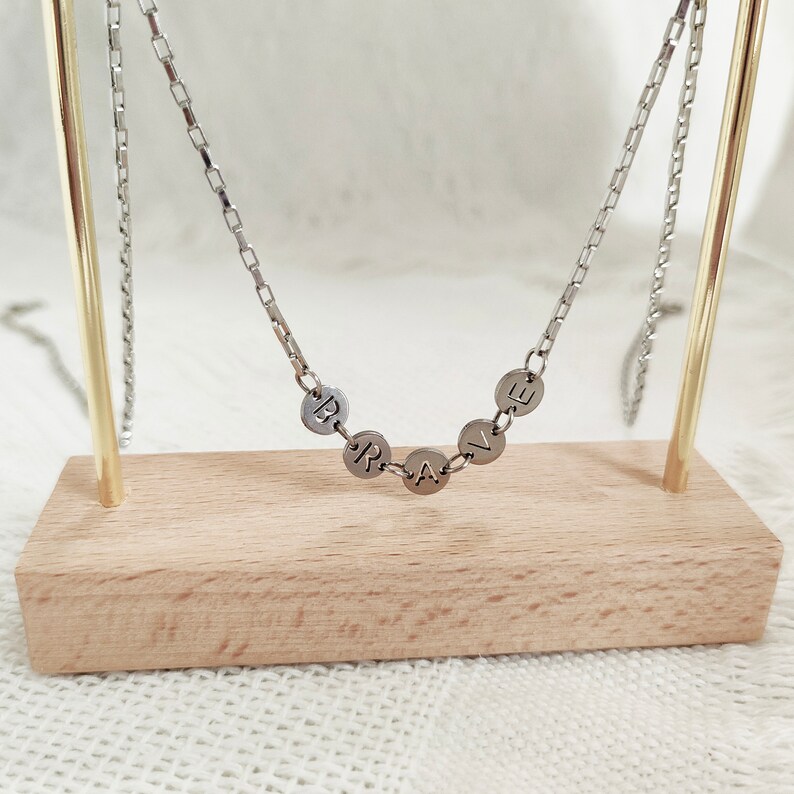 May include: A silver chain necklace with a pendant that spells out the word "BRAVE" in individual silver circles.