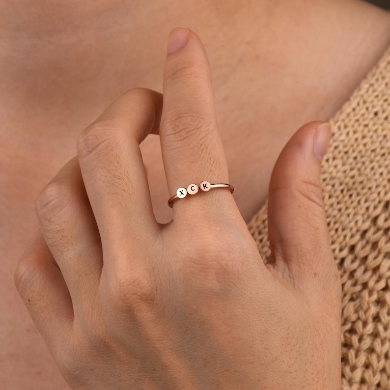 May include: A rose gold ring with three small round charms, each with a letter engraved on it. The letters are X, C, and K.