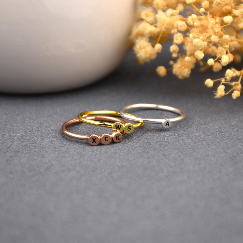 May include: Three delicate rings with initial charms. The rings are stacked on a grey surface. The top ring is silver with the letter "A" charm. The middle ring is gold with the letters "W" and "S" charms. The bottom ring is rose gold with the letters "X", "C", "K" charms.