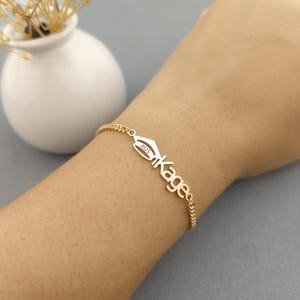 Puede incluir: Pulsera dorada con un dije de birrete de graduación y el nombre "Kagee" en fuente cursiva. El dije incluye el año "2025". La pulsera se lleva en la muñeca.