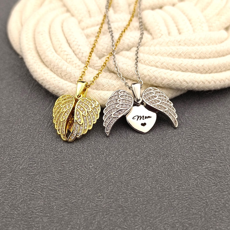 May include: Two silver necklaces with angel wing pendants. One pendant is a heart with the word "Mom" inscribed on it. The other pendant is a pair of angel wings.