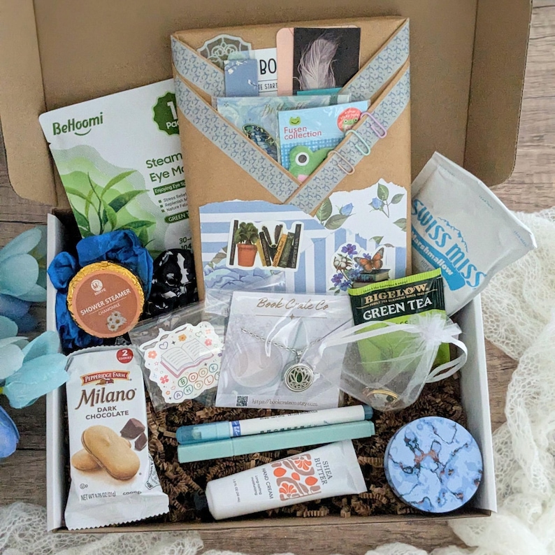May include: A gift box filled with self-care items. Contents include a steam eye mask, Milano dark chocolate cookies, a necklace, Bigelow green tea, a Swiss Miss marshmallow packet, and other small gifts. The box is decorated with paper and stickers.