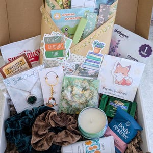 May include: A gift box filled with various items, including a necklace, scrunchies, a candle, tea, a bookmark, and stickers. The box contains a variety of colors and textures, creating a cozy and inviting aesthetic. The theme appears to be relaxation and self-care.