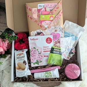 May include: A gift box filled with self-care items. Includes a steam eye mask, Milano dark chocolate cookies, green tea, hand cream, a scrunchie, and stickers. The box is decorated with pink and floral accents.