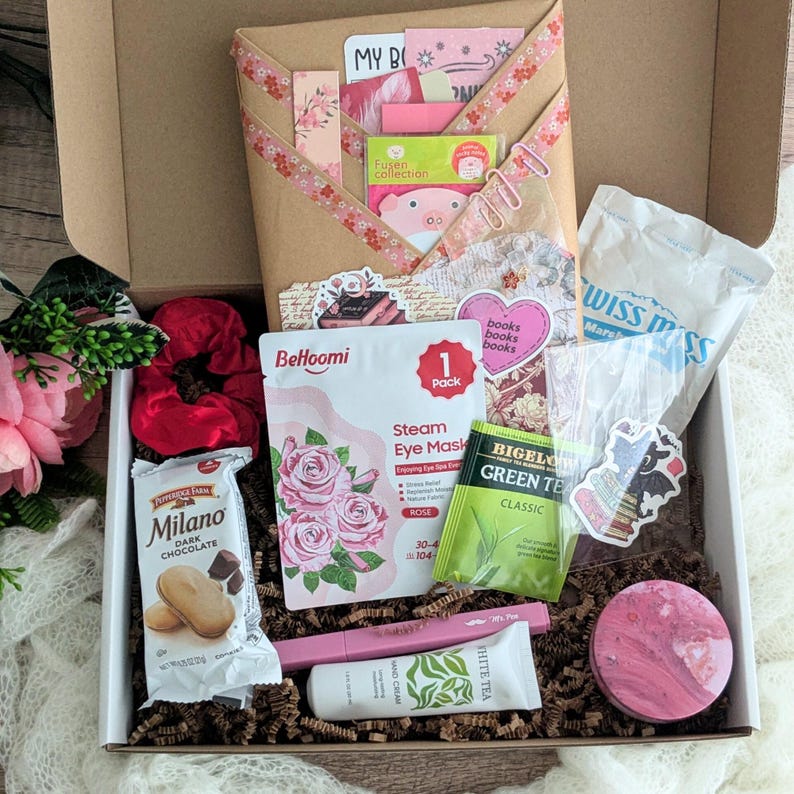 May include: A gift box filled with self-care items. Includes a steam eye mask, Milano dark chocolate cookies, green tea, hand cream, a scrunchie, and stickers. The box is decorated with pink and floral accents.