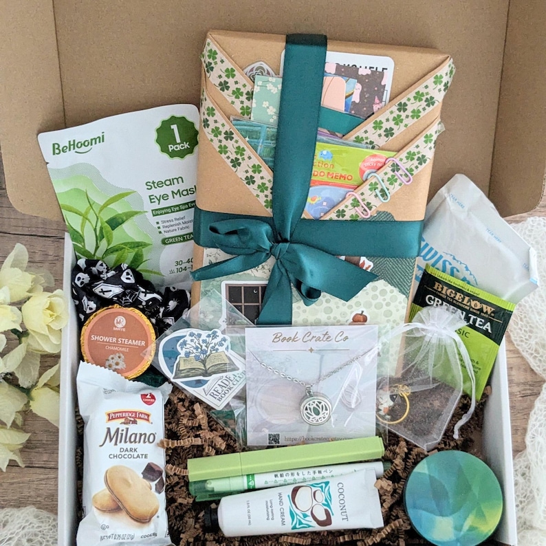 May include: A gift box filled with various items, including a wrapped package with a green ribbon, a necklace, tea bags, eye masks, cookies, and hand cream. The box contains a variety of self-care and relaxation products.