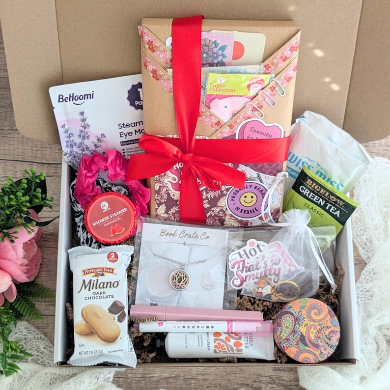 May include: A gift box filled with various items, including a wrapped package tied with a red ribbon, a necklace, Milano cookies, tea bags, and beauty products. The box contains a variety of colors and textures, creating a visually appealing arrangement.
