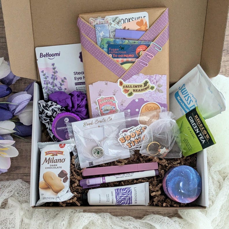 May include: A gift box filled with various items, including a necklace, hand cream, tea, and snacks. The box contains a decorative envelope with the text "FALL INTO READING" and "BOOKISH" stickers. The color scheme is primarily purple and white.