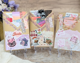 Op de afbeelding: Drie decoratieve enveloppen met boeken als thema op transparante standaards. Elke envelop is versierd met stickers, papier met patronen en versieringen. De stickers tonen zinnen als "Books", "I Read" en "Book Lover", samen met bloemen- en boekillustraties.