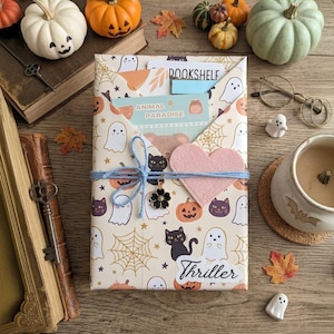 May include: A rectangular package wrapped in cream-coloured paper featuring a pattern of ghosts, black cats, pumpkins, and spiderwebs. It is tied with blue twine, a pink felt heart, and a black flower charm. A sticker labelled Thriller is attached.