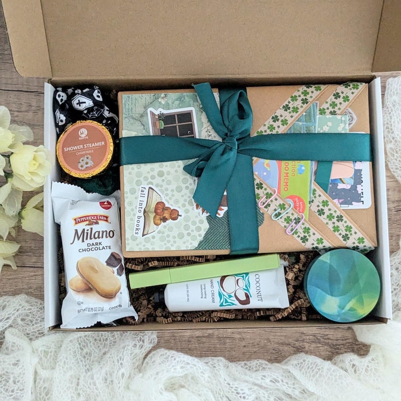 May include: A gift box filled with various items, including a wrapped package tied with a teal ribbon, a package of Milano dark chocolate cookies, a shower steamer, hand cream, and a decorative round object. The box is open, revealing the contents.